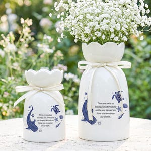 May include: Two white ceramic vases with scalloped rims, each tied with a cream ribbon. The vases feature blue illustrations of a whale, a sea turtle, a starfish, and a seashell, along with the text: There are souls as beautiful and formidable as the sea; blessed are those who encounter one of them.