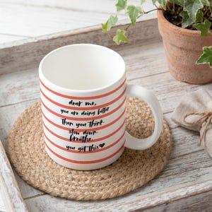 Könnte beinhalten: Weiße Keramiktasse mit roten horizontalen Streifen und einem gebogenen Henkel. Auf der Tasse steht der Text "dear me, you are doing better than you think. slow down. breathe. you got this" in Schwarz. Die Tasse steht auf einem runden, gewebten Untersetzer.