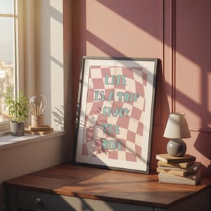 Könnte beinhalten: Ein gerahmter Kunstdruck mit dem Text "LIFE IS A TRIP ENJOY THE RIDE" in einem rosa-cremefarbenen Schachbrettmuster. Der Druck steht auf einem Holztisch, neben einer kleinen Lampe und einem Stapel Bücher.