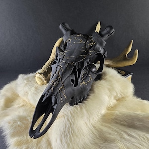 May include: A black decorative deer skull with gold crackle detailing, resting on a white faux fur throw. The skull has large eye sockets and prominent antlers. The background is a dark gray.