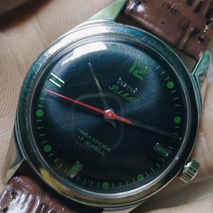 May include: A vintage Pilot wristwatch with a dark dial, green numerals, and a red second hand. The watch has a silver-colored case and a brown leather strap. The dial reads "PARA SHOCK 17 JEWELS".