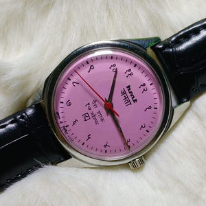 May include: A vintage wristwatch with a pink face and black leather band. The watch face features Arabic numerals and text in a foreign language. The watch hands are black and red, and the watch is set against a white, fluffy background.