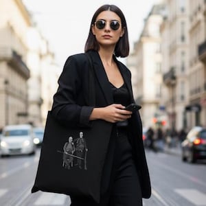 Puede incluir: Un bolso tote negro con una ilustración en blanco y negro de dos figuras, llevado por una persona con blazer, pantalón y mocasines negros. La persona camina por una calle adoquinada.