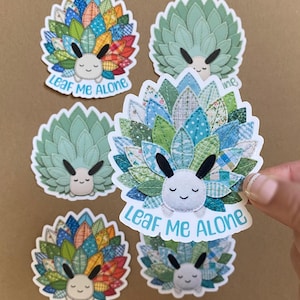 May include: A collection of whimsical stickers featuring a smiling, white-faced creature with leaf-like details. The leaves are a mix of colors and patterns, with the text "Leaf Me Alone" in a playful font. The stickers are on a neutral background.