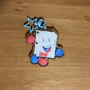 Op de afbeelding: Een heldere acryl sleutelhanger met een cartoontekening van een lachend stuk papier met een rode vlinderdas, rode en blauwe armen en benen, en een blauwe stervormige sluiting. De sleutelhanger ligt op een houten oppervlak.