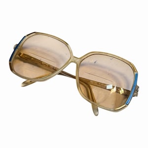 Könnte beinhalten: Vintage übergroße Sonnenbrille mit leicht brauner Tönung. Die Fassungen sind hellbeige mit blauen Akzenten an den Bügeln. Der Text "CAPRI VIENNA" ist auf dem Glas sichtbar.