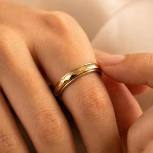 May include: A gold band ring featuring a faceted, diamond-cut texture along the center. The ring has a polished finish and a slim profile, showcasing a repeating geometric pattern that reflects light across its metallic surface.