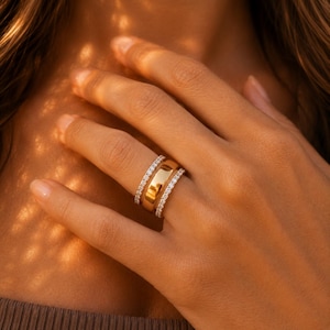 Puede incluir: Un conjunto de tres anillos en un dedo. El anillo central es una banda ancha de oro pulido con una superficie lisa y reflectante. Está flanqueado a ambos lados por dos anillos de eternidad más finos a juego, engastados con pequeñas piedras claras.