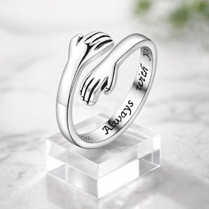 May include: A silver-colored ring with an open design, featuring two hands embracing. The ring is engraved with the words "Always with you". The ring is displayed on a clear, square stand.
