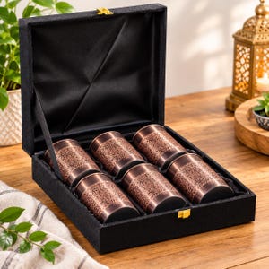 May include: A black velvet-lined presentation box containing six cylindrical copper-toned metal tumblers. Each tumbler features an intricate embossed floral pattern around the exterior. The box has a hinged lid with a gold-coloured metal latch.