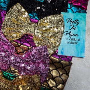 May include: An assortment of sequined hair bows in various colors, including gold, black, purple, and teal. The bows have a mermaid scale pattern and are handmade. A card with the text "Pretty In Aqua Personalized Handmade" is also present.