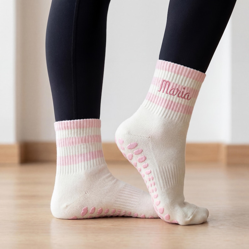 May include: Cream-colored socks with pink stripes and the name "Maria" embroidered on the side. The socks have pink grip dots on the soles. The person is wearing black leggings.