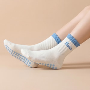May include: A pair of white socks with blue accents. The socks have a ribbed texture and a blue cuff. The word "Maria" is embroidered in blue on the side of one sock. The soles of the socks have a blue floral pattern.