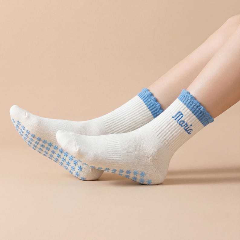 May include: A pair of white socks with blue accents. The socks have a ribbed texture and a blue cuff. The word "Maria" is embroidered in blue on the side of one sock. The soles of the socks have a blue floral pattern.