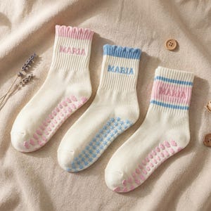 May include: Three pairs of cream-colored socks with the name "MARIA" embroidered on the cuffs. Each pair has a different color cuff: pink, blue, and pink with blue stripes. The soles have pink non-slip grips.