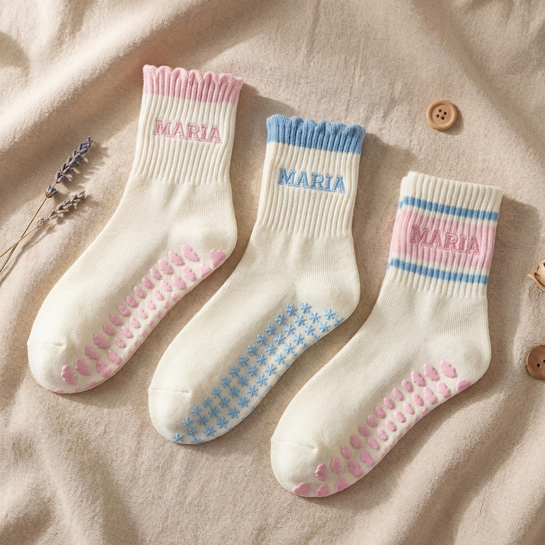 May include: Three pairs of cream-colored socks with the name "MARIA" embroidered on the cuffs. Each pair has a different color cuff: pink, blue, and pink with blue stripes. The soles have pink non-slip grips.