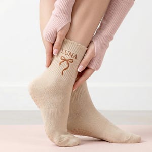 Può includere: Un paio di calzini beige con bordo smerlato. La parola "LUNA" e un fiocco sono ricamati in marrone sul bordo. I calzini hanno un'aderenza antiscivolo sulle suole. La persona indossa scaldabraccia rosa.