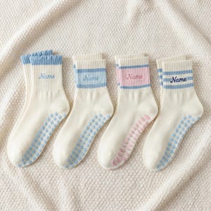 May include: Four pairs of white socks with blue or pink accents. Each sock has the word "Name" embroidered on the cuff. The socks have a floral pattern on the soles and stripes on the cuffs. The cuffs are blue or pink.