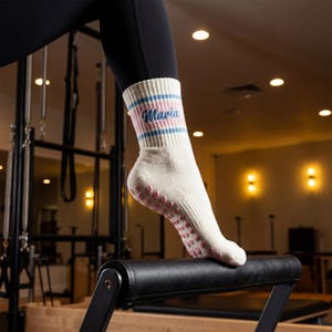 May include: Cream-colored athletic socks with blue and pink stripes and the name "Maria" embroidered on the cuff. The socks have pink non-slip grips on the sole. The person is wearing black leggings.