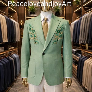May include: A sage green blazer featuring intricate floral embroidery on the chest, worn over a white shirt with a gold textured tie. The blazer has a single button closure and two patch pockets. The mannequin has a gold head and gold hands.