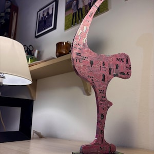 Può includere: Una scultura astratta rosa con un design a stampa di giornale. La scultura ha una forma alta e curva e poggia su una base nera con accenti verdi. La scultura è su una scrivania di legno.