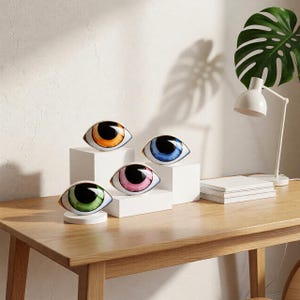 Puede incluir: Cuatro objetos decorativos de cerámica con forma de ojo, con iris de diferentes colores: naranja, azul, rosa y verde. Cada ojo tiene una pupila negra y un borde dorado. Se exhiben sobre pedestales blancos en una mesa de madera.