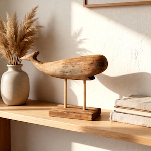 Peut inclure: Sculpture en bois représentant une baleine, avec une finition naturelle, montée sur une base en bois avec deux fines pattes. La baleine est exposée sur une étagère en bois clair, à côté d'un vase avec de l'herbe de la pampa séchée et des draps pliés de tons neutres.