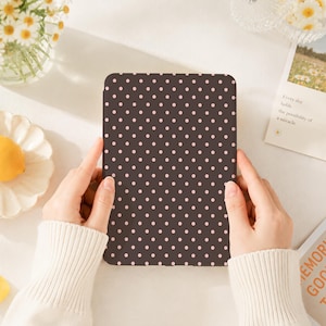 May include: A rectangular electronic e-reader device featuring a dark brown cover with a repeating pattern of small, light pink polka dots. The device is held upright, displaying a uniform grid of dots across its entire front-facing surface area.