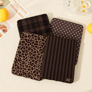 May include: Four rectangular e-reader cases featuring distinct patterns: a dark brown plaid, a dark brown with pink polka dots, a tan and black leopard print, and a dark brown vertical stripe. The leopard and striped cases feature the gold letters RC.