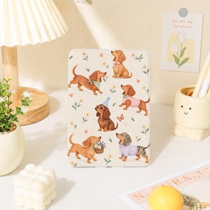 May include: A rectangular tablet case featuring a cream-coloured background with a repeating pattern of brown dachshund dogs. The dogs are depicted in various poses, wearing accessories like a blue party hat, sunglasses, a pink bow, and sweaters.
