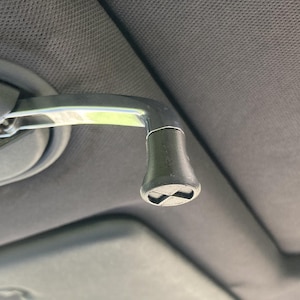 May include: A black, textured, flared handle knob attached to a metallic car interior grab handle. The knob features a flat, circular end cap with a recessed cross-shaped design. The handle is mounted against a dark gray, fabric-covered vehicle ceiling.