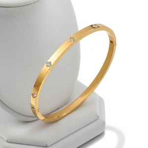 May include: A gold-colored bangle bracelet with four small, clear gemstones. The bracelet is displayed on a white stand against a white background. The bracelet has a simple, elegant design.