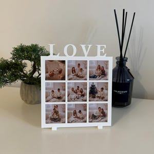 Könnte beinhalten: Weißer Fotorahmen mit neun Öffnungen, die jeweils ein Familienfoto enthalten. Das Wort "LOVE" ist in weißen Buchstaben über dem Rahmen geschrieben. Eine kleine Topfpflanze und eine schwarze Diffusorflasche befinden sich auf beiden Seiten des Rahmens.