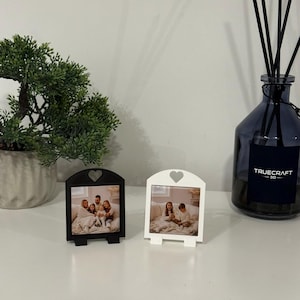 May include: Two small photo frames, one black and one white, each with a heart-shaped cutout at the top. The frames display family photos and stand on small easel-like supports. A bonsai tree and a blue glass diffuser bottle are in the background.