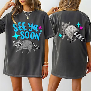 Può includere: T-shirt grigio scuro con grafica di procione e la scritta "SEE YA SOON" in blu. Il procione è grigio e bianco con la coda a strisce. Il design include stelle blu e rosa.