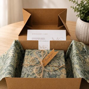 An open brown cardboard gift box containing a package wrapped in patterned green floral paper, tied with twine and a small brown tag.