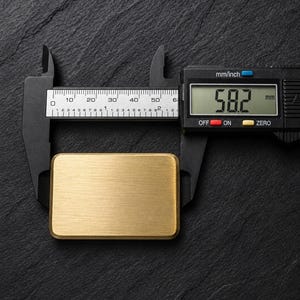 A rectangular solid brass plaque placed horizontally on a dark slate surface, being measured with black digital calipers showing a length of 58.2 millimeters.