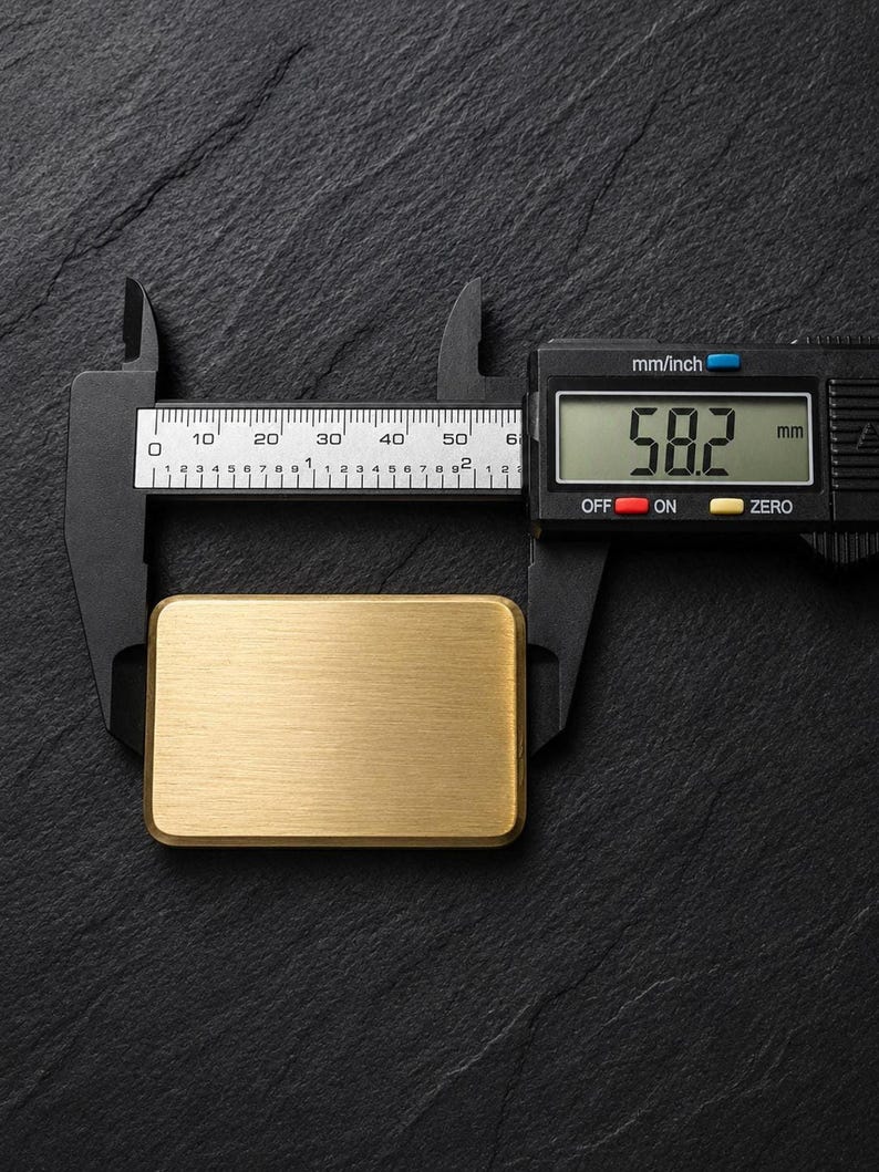A rectangular solid brass plaque placed horizontally on a dark slate surface, being measured with black digital calipers showing a length of 58.2 millimeters.