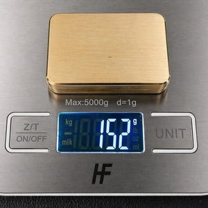 A rectangular solid brass plaque rests on a digital scale showing a weight of 152 grams.