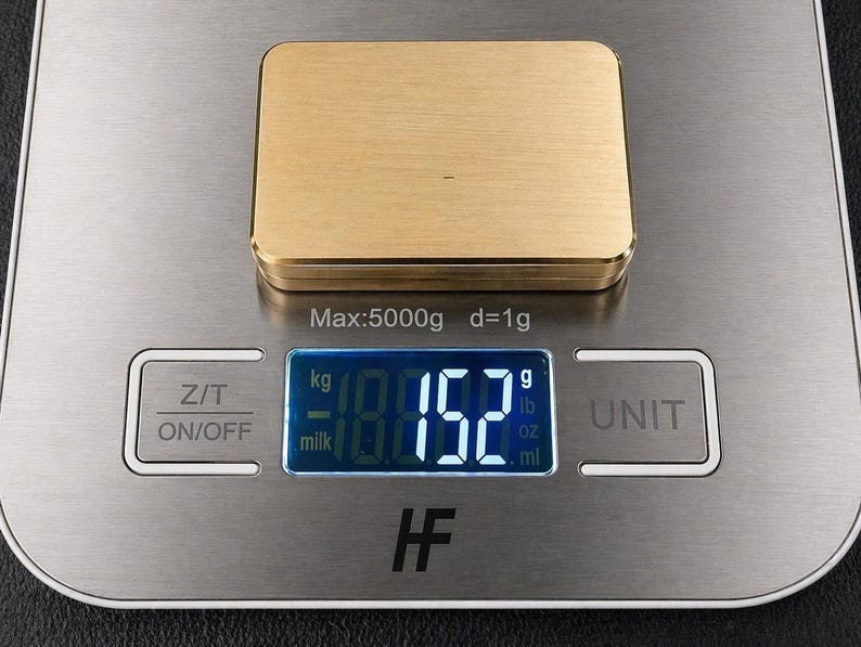 A rectangular solid brass plaque rests on a digital scale showing a weight of 152 grams.