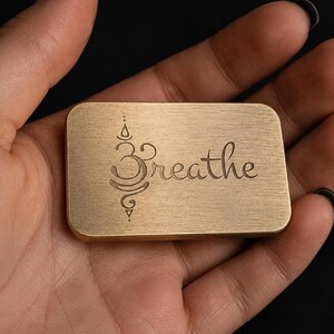 A hand holds a rectangular solid brass token engraved with the word breathe and a lotus motif.