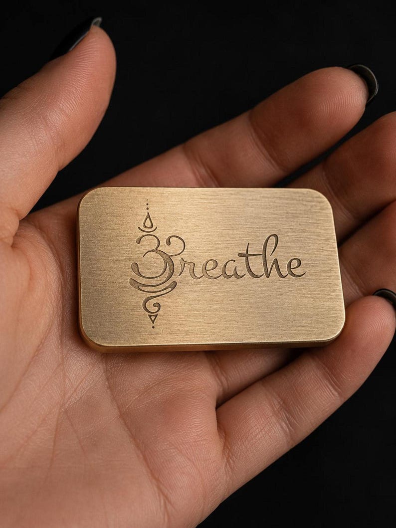 A hand holds a rectangular solid brass token engraved with the word breathe and a lotus motif.