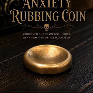 Op de afbeelding: Een ronde, goudkleurige messing wrijfmunt met een glad, concaaf midden op het bovenoppervlak. De tekst ANXIETY RUBBING COIN staat erboven, met aanvullende tekst die CONCAVED SHAPE ON BOTH SIDES, REAR SIDE CAN BE PERSONALISED en SOLID BRASS vermeldt.