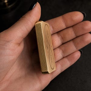 A hand holds the side profile of a rectangular solid brass breathing anchor token.