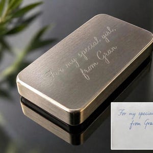 A solid brass plaque engraved with the cursive text For my special girl, from Gran sits on a dark reflective surface next to an inset image of the original handwritten note.