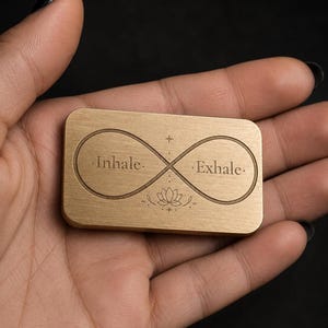 A hand holds a rectangular solid brass token engraved with an infinity symbol, lotus, and the words inhale and exhale.