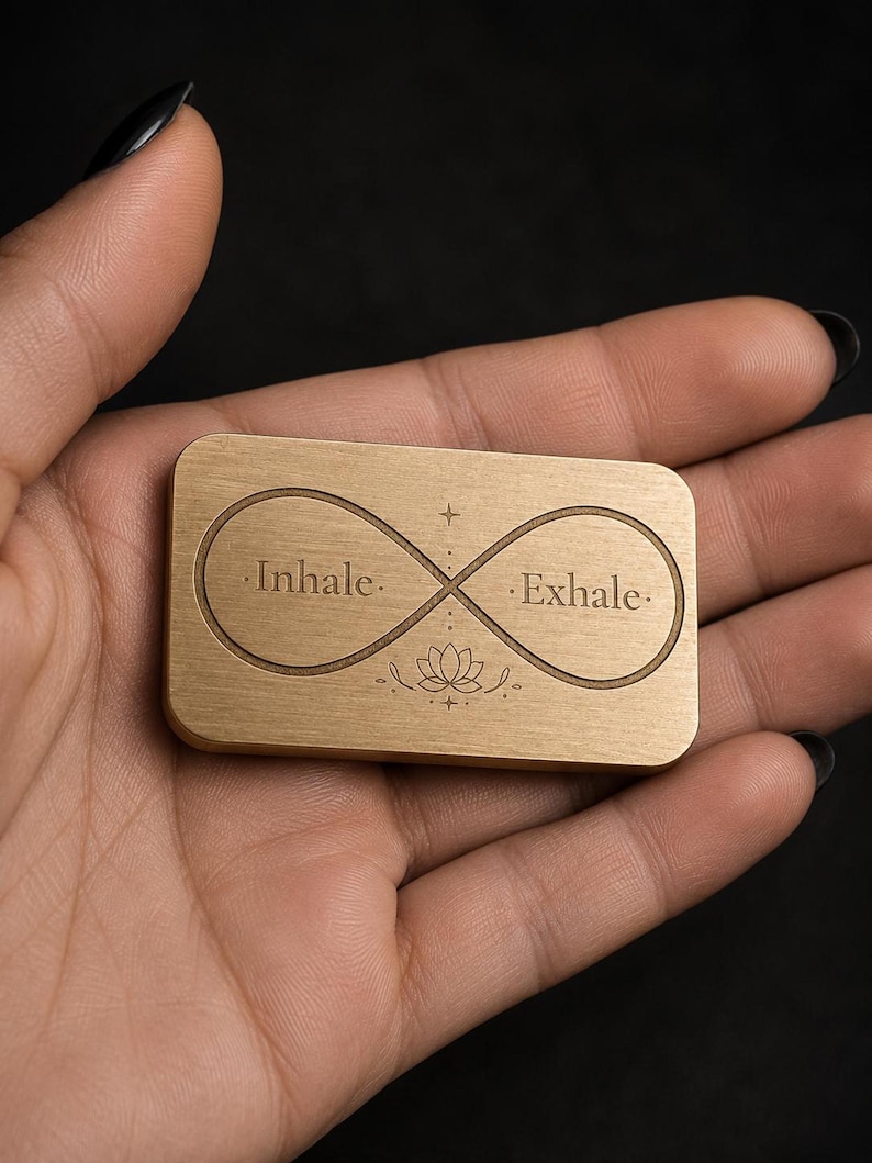 A hand holds a rectangular solid brass token engraved with an infinity symbol, lotus, and the words inhale and exhale.
