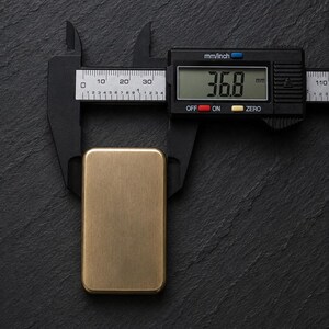 A rectangular solid brass plaque placed vertically on a dark slate surface, being measured with black digital calipers showing a width of 36.8 millimeters.