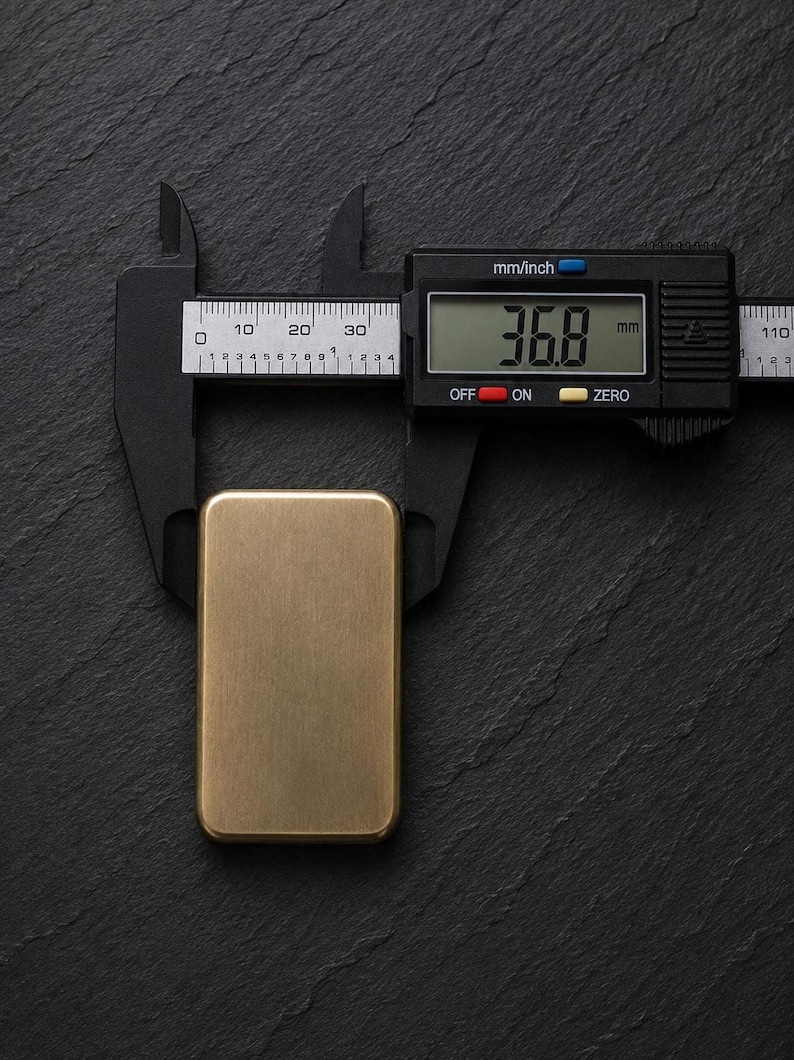 A rectangular solid brass plaque placed vertically on a dark slate surface, being measured with black digital calipers showing a width of 36.8 millimeters.