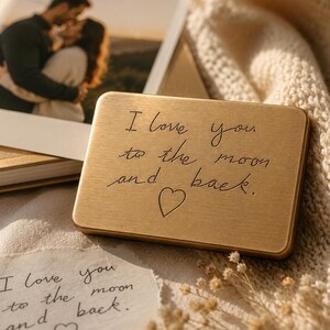 An engraved solid brass plaque with the words I love you to the moon and back rests on a knitted fabric surface next to a framed photo of a couple and a handwritten paper note.